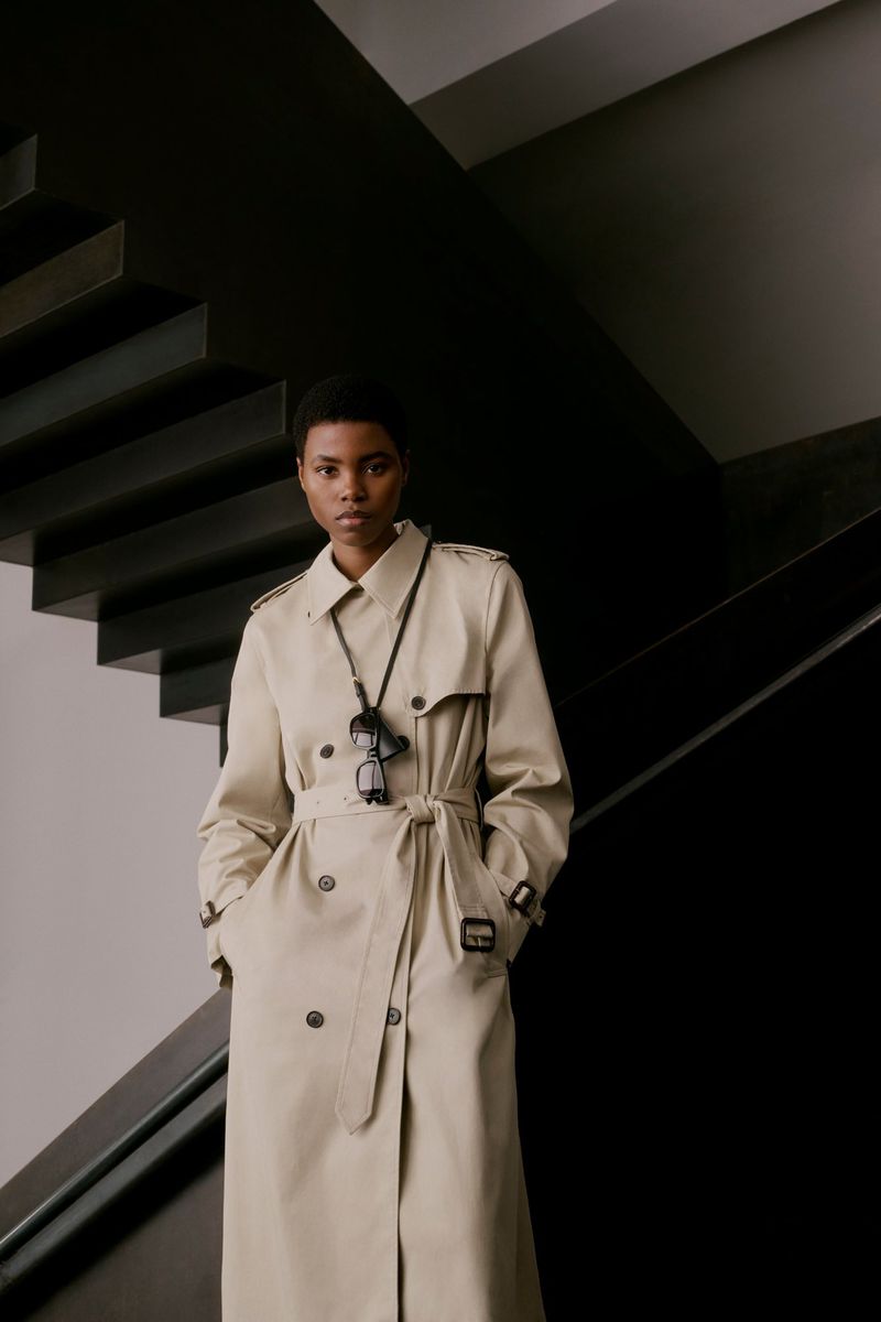 A Black woman in a light trench coat stands on black stairs with a modern aesthetic, complemented by her stylish eyewear.
