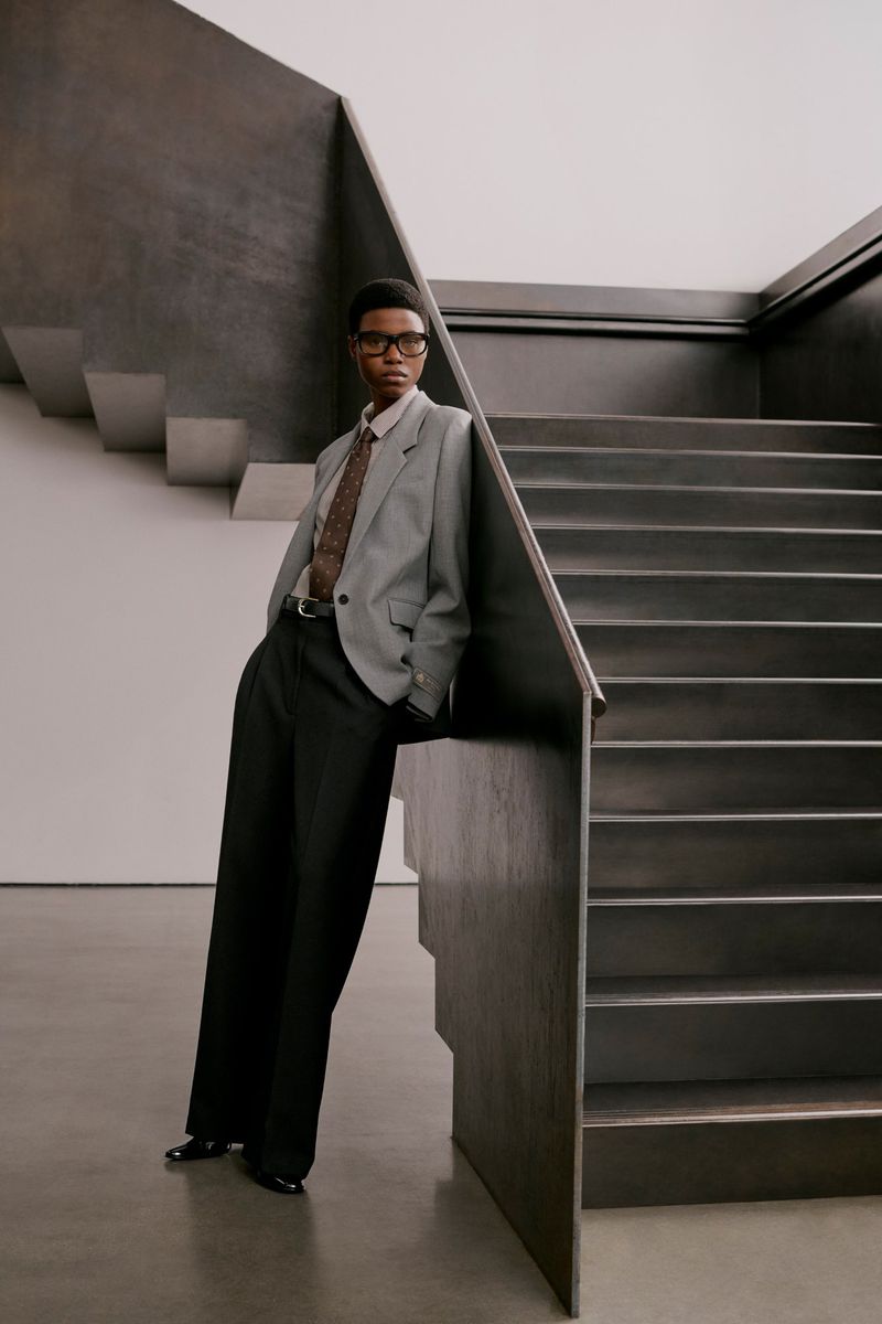 Model wearing a grey blazer and black trousers leaning against a dark staircase for Aritzia