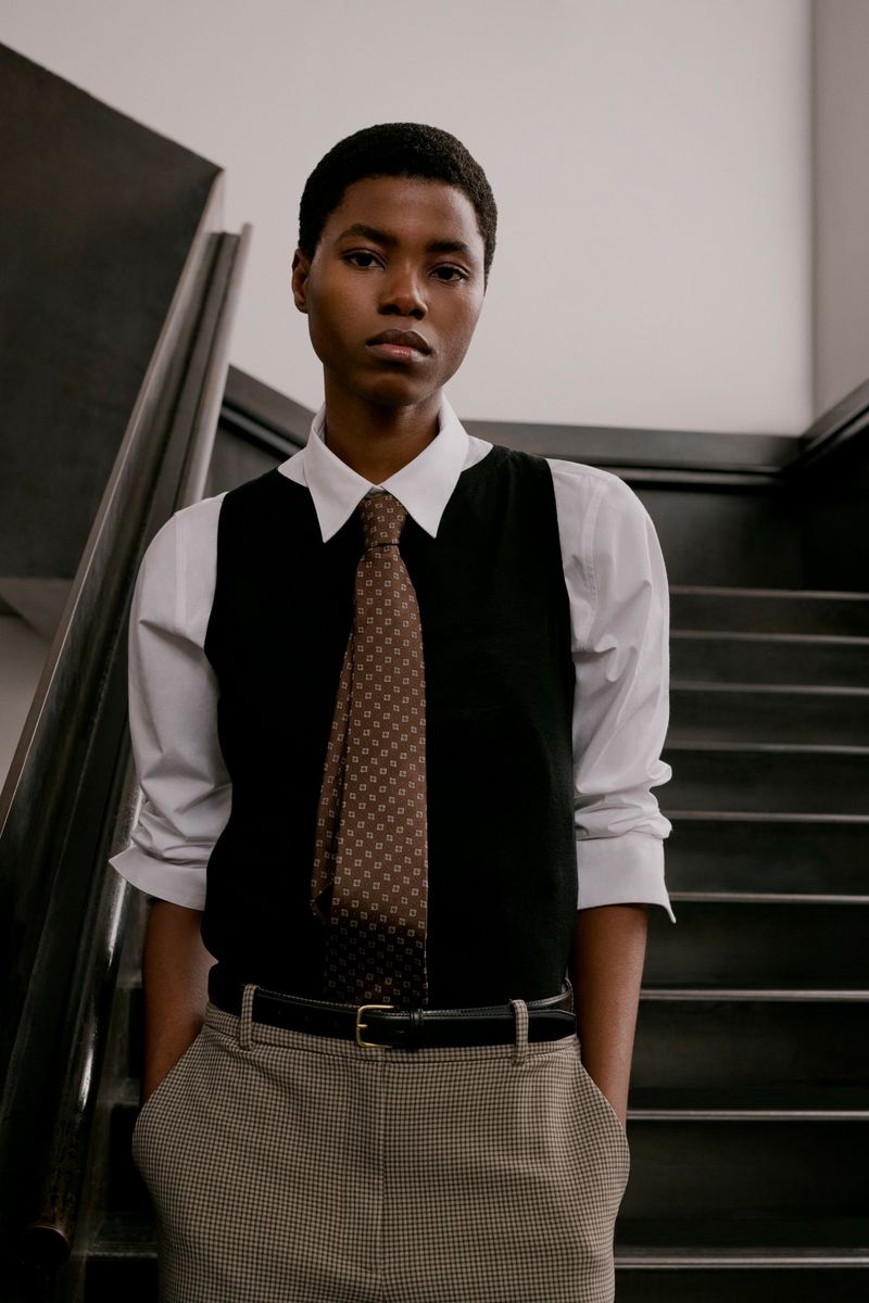 Model wearing a white collared shirt, black vest, patterned tie, and trousers standing on a staircase