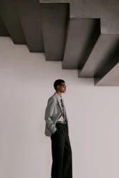 Model wearing a grey blazer and black trousers standing below sculptural concrete stairs against a plain wall