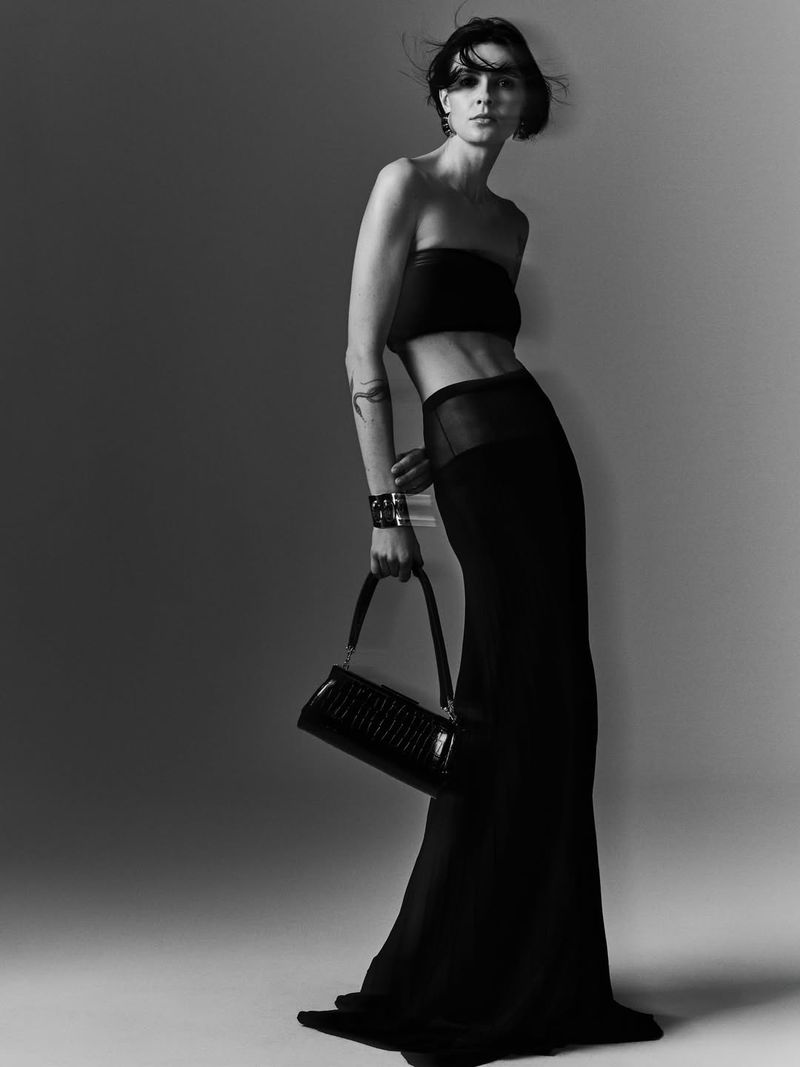 Model in a black bandeau top and long skirt holding a black handbag against a grey studio backdrop.