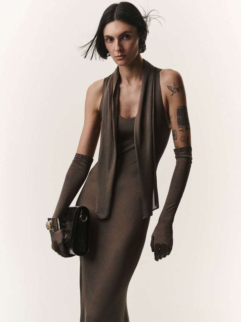 Model in brown dress and long gloves holding a black handbag against white seamless