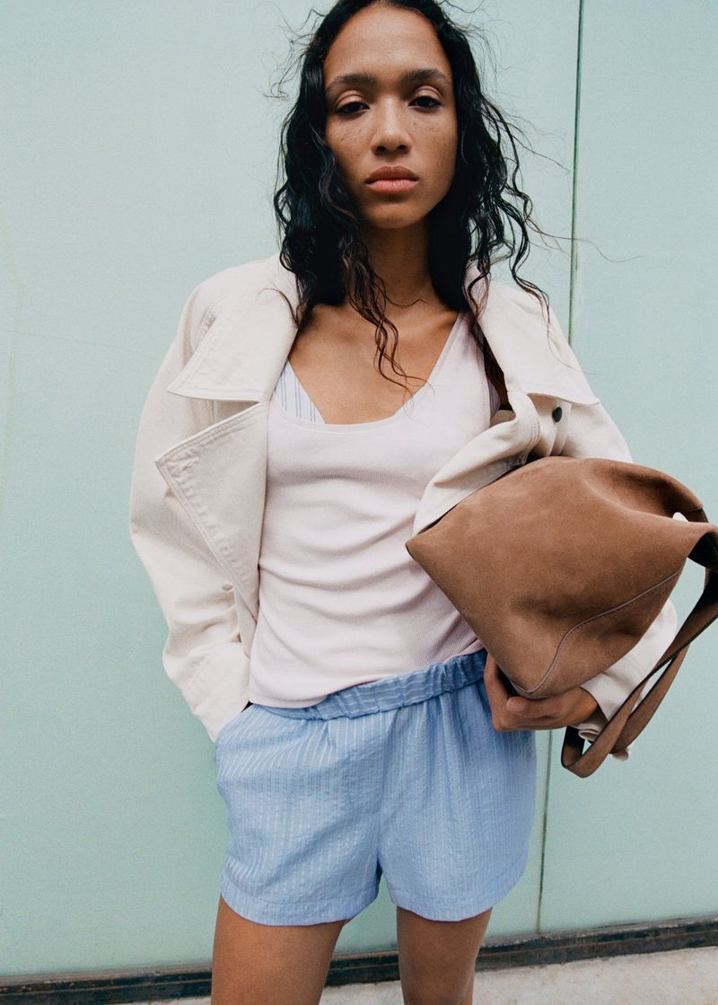 Model wearing a light jacket, white top, and blue shorts holding a brown suede bag against a pale turquoise wall.
