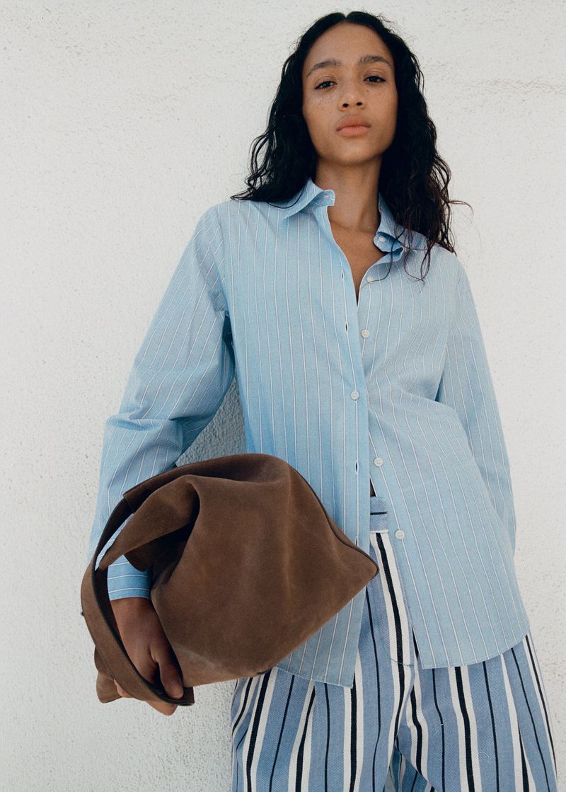 Model wearing a light blue striped button-down shirt and holding a brown suede handbag against a white textured wall.