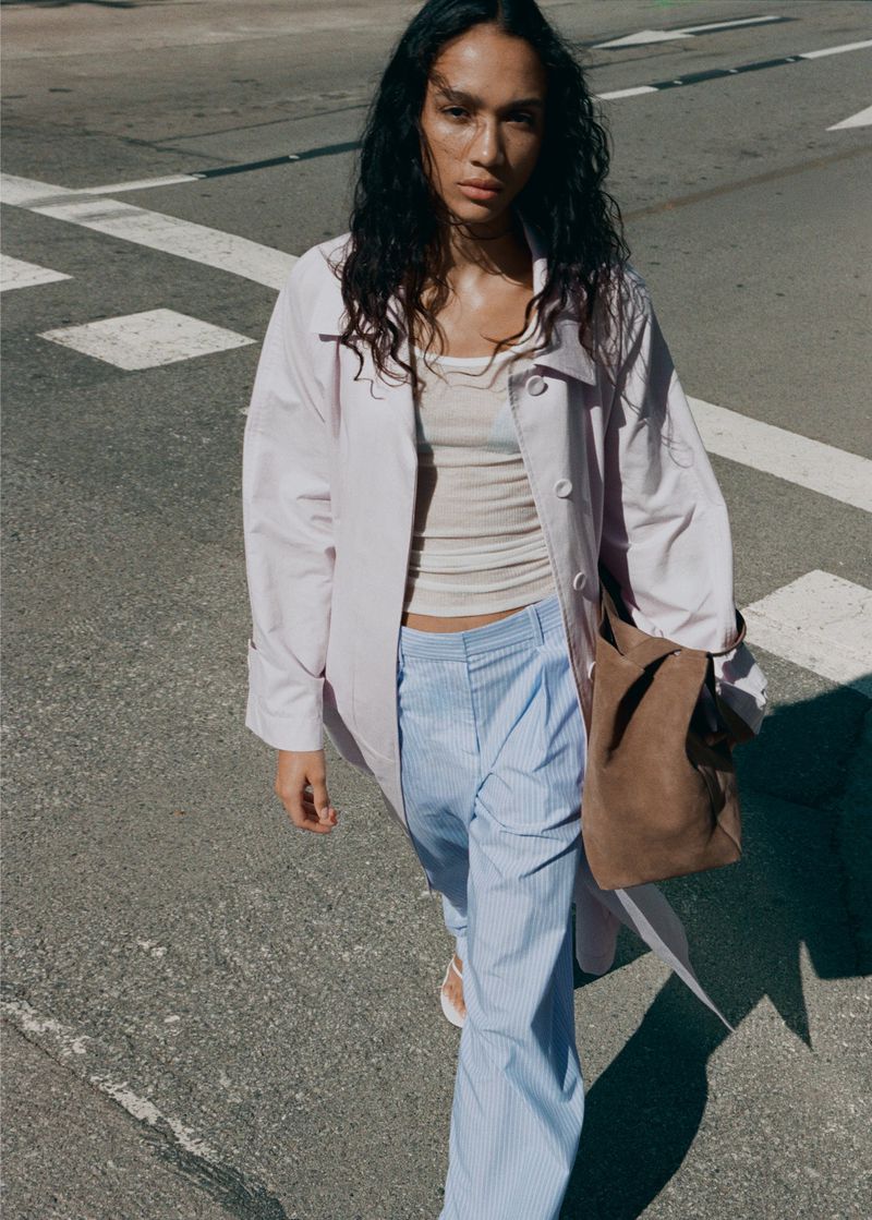 Model walking on asphalt street wearing light pink coat, striped trousers and holding brown suede handbag