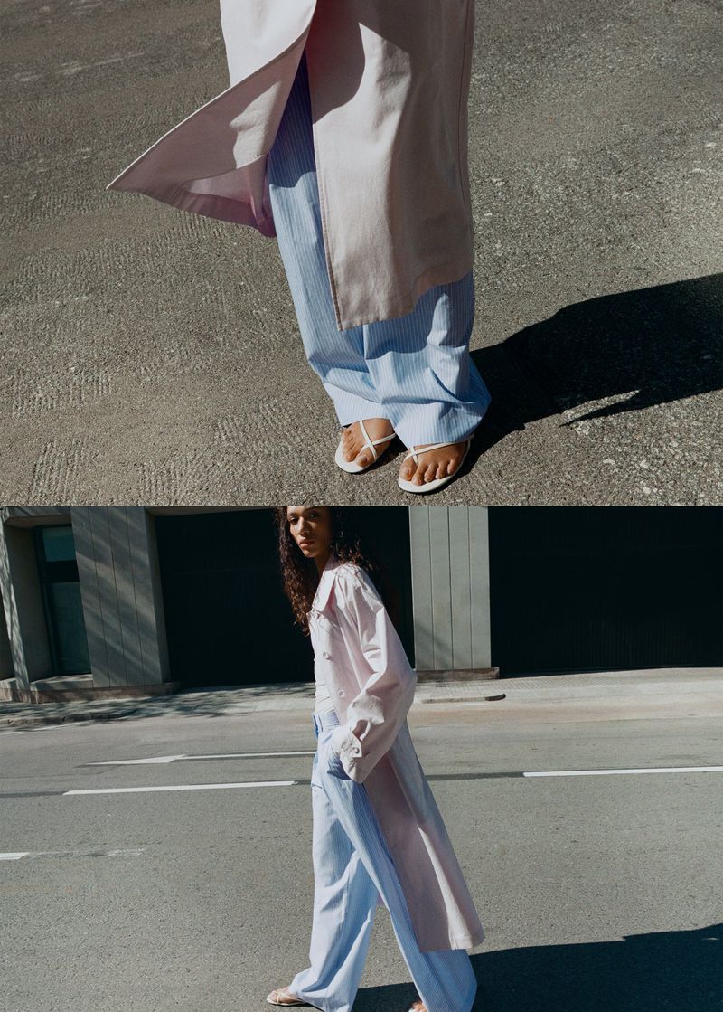 Model wearing a pale pink coat and light blue striped trousers standing on an asphalt street with white pavement markings.