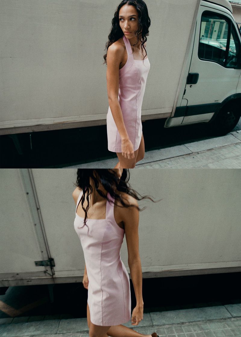 A young woman with long, dark, wavy hair walks on a city street next to a truck, wearing a light pink mini dress and sandals.