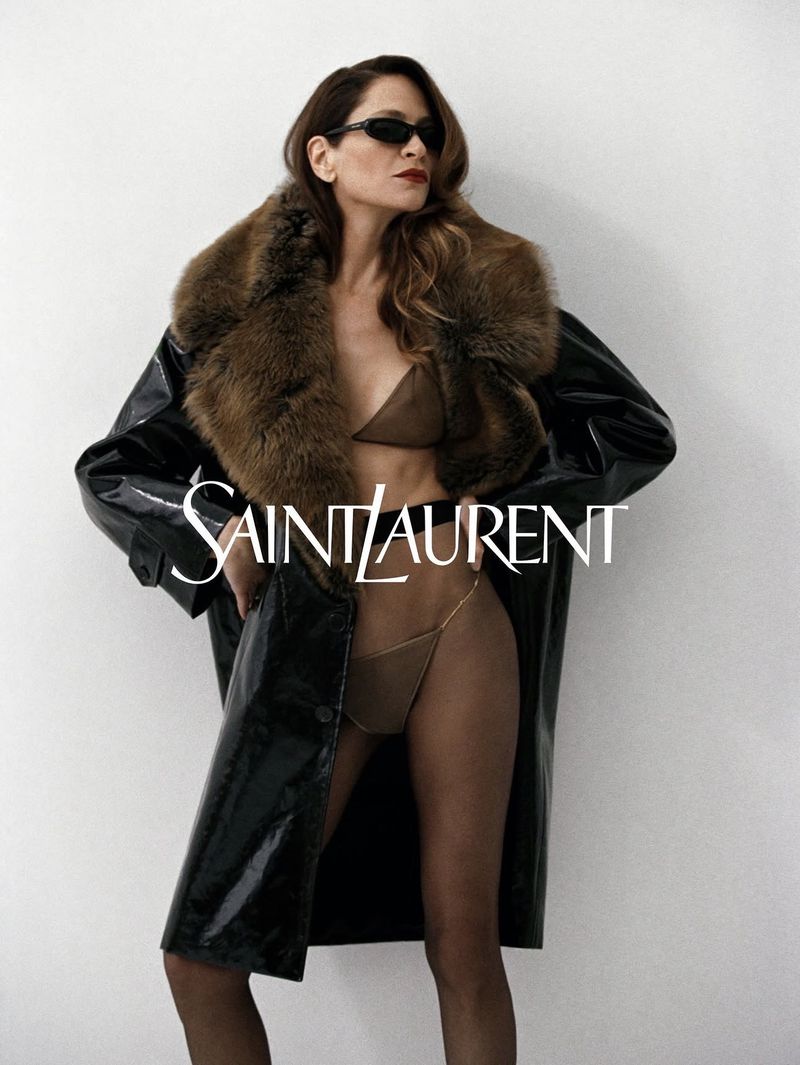 Model Frankie Rayder wearing a black patent coat with fur collar and dark sunglasses against a white background