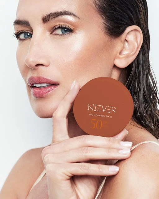 Beauty shot of an adult woman holding a brown compact; the makeup product, 'NIEVES daily skin protector SPF 50,' is clearly displayed.