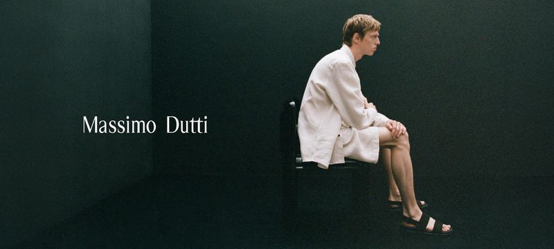 An adult male in a cream linen suit sits in a dark studio. The man is facing right in sandals with the brand name Massimo Dutti on the left.
