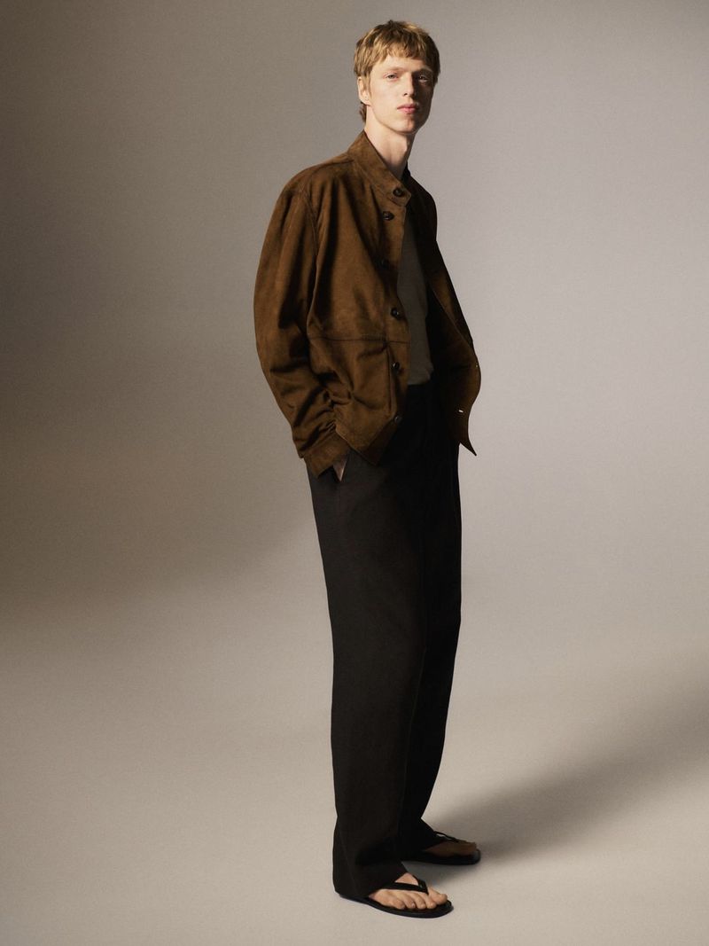 A fair-skinned male model in a brown suede jacket and black slacks stands in a neutral studio setting with a soft, diffused lighting style.