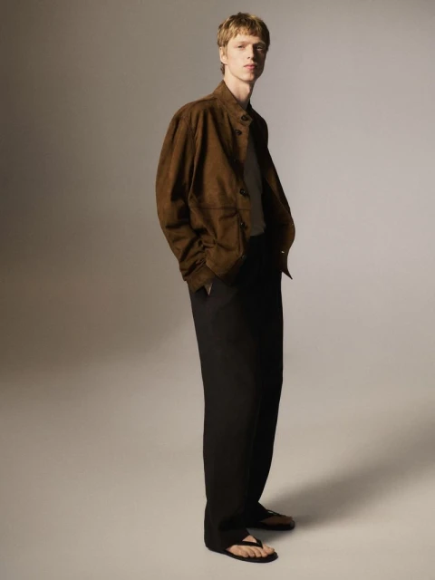 A fair-skinned male model in a brown suede jacket and black slacks stands in a neutral studio setting with a soft, diffused lighting style.