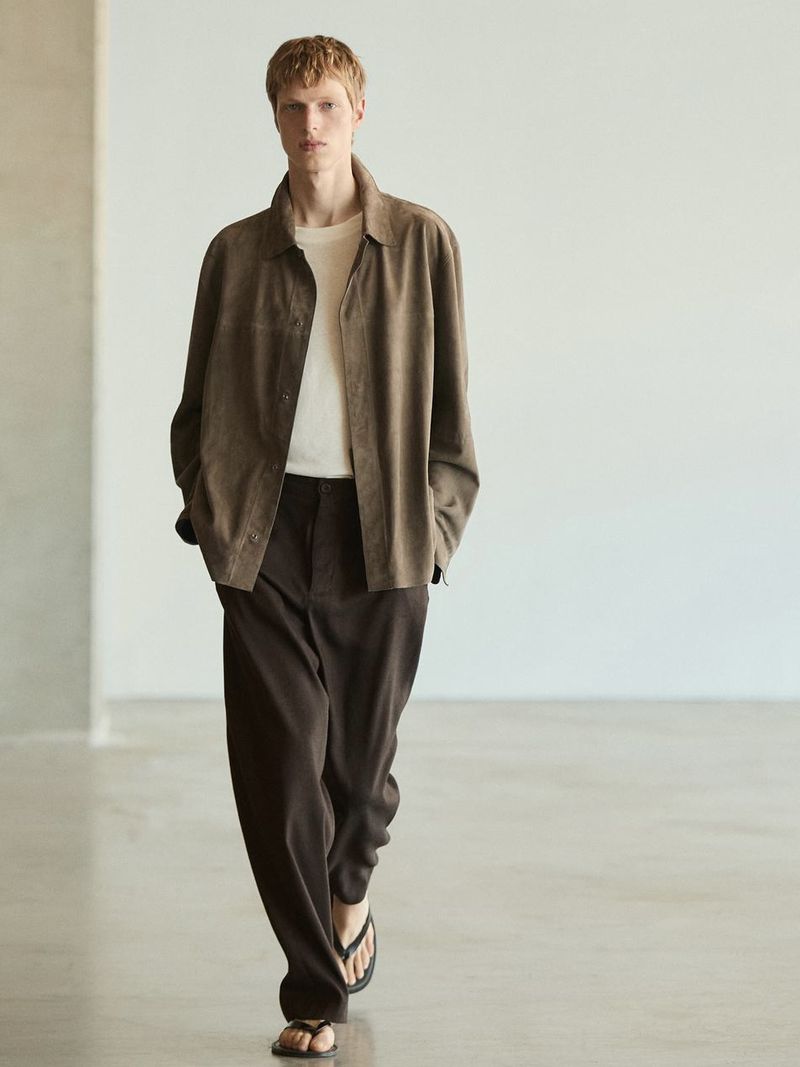 A blonde male model walks in brown jacket, cream t-shirt, and brown pants, with sandals, posing against a neutral wall in a fashion editorial.