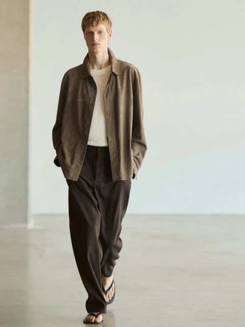 A blonde male model walks in brown jacket, cream t-shirt, and brown pants, with sandals, posing against a neutral wall in a fashion editorial.