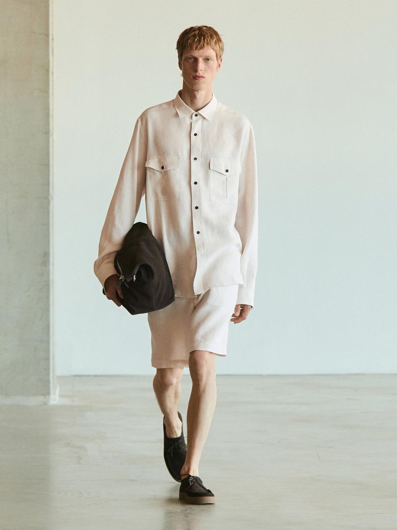 A stylish adult male walks in a photo studio wearing a light-pink long-sleeve shirt, matching shorts, and black shoes, carrying a black bag.
