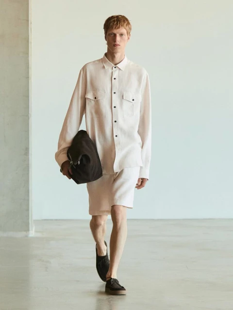 A stylish adult male walks in a photo studio wearing a light-pink long-sleeve shirt, matching shorts, and black shoes, carrying a black bag.