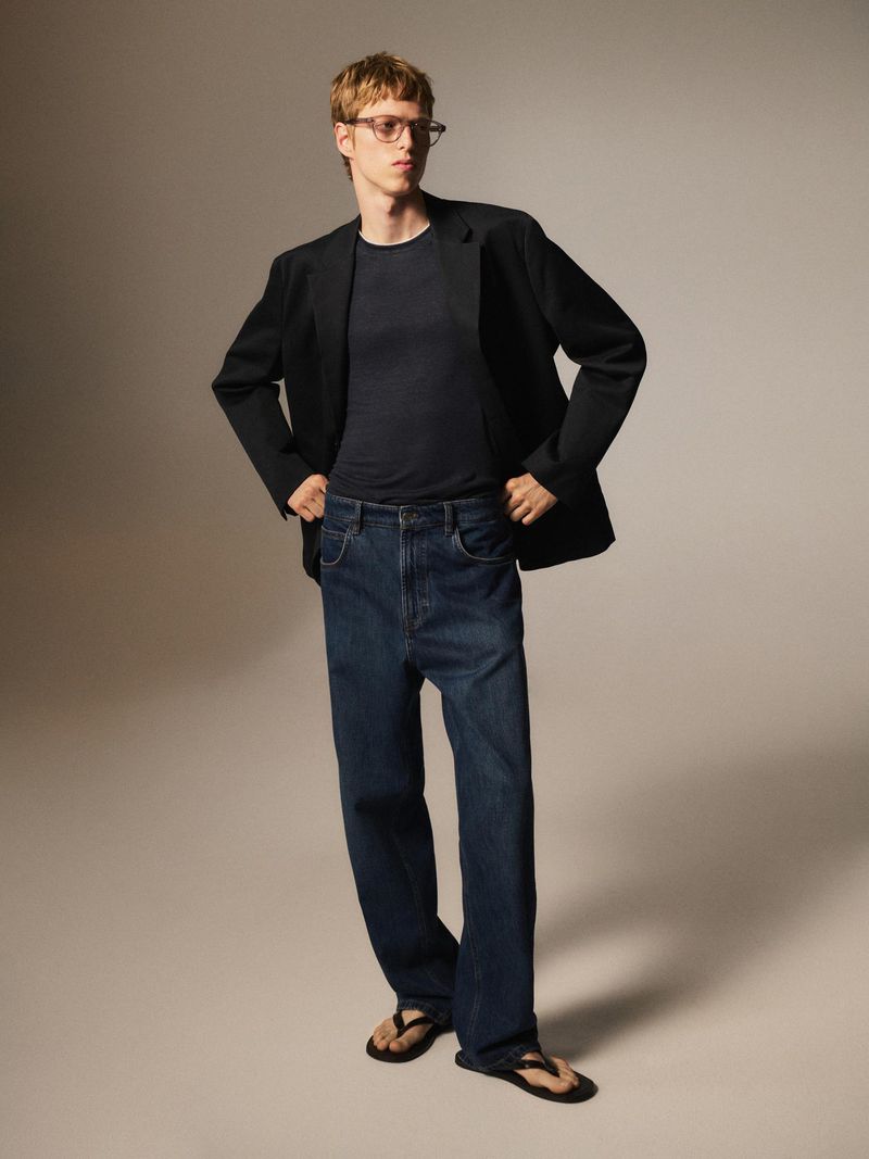 Man in a studio setting wearing a black blazer, dark blue jeans and thong sandals standing with his hands on his hips.