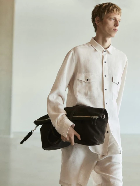 Fashion shot of an adult male wearing a collared shirt and shorts, carrying a brown handbag in a brightly lit studio setting.