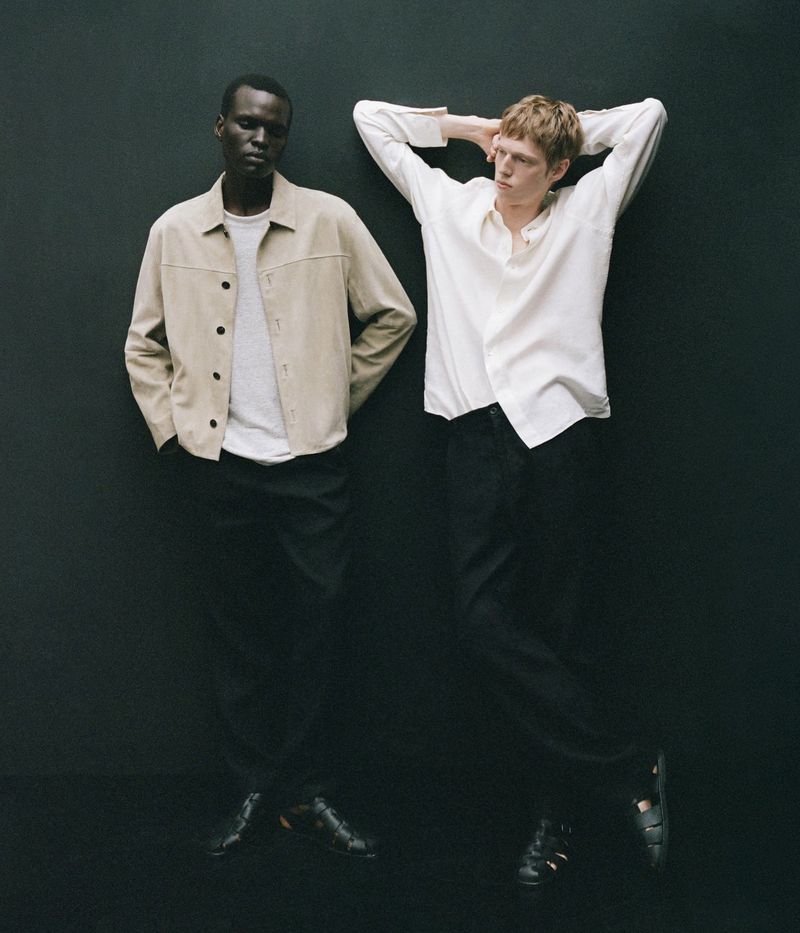 Two stylish adult men in a fashion studio. One Black man stands next to a fair-skinned man with neutral expressions, wearing casual outfits.
