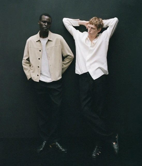 Two stylish adult men in a fashion studio. One Black man stands next to a fair-skinned man with neutral expressions, wearing casual outfits.