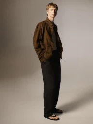 Model Joseph Uyttenhove poses in a brown suede jacket, wide-leg black trousers, and leather sandals against a neutral studio backdrop.