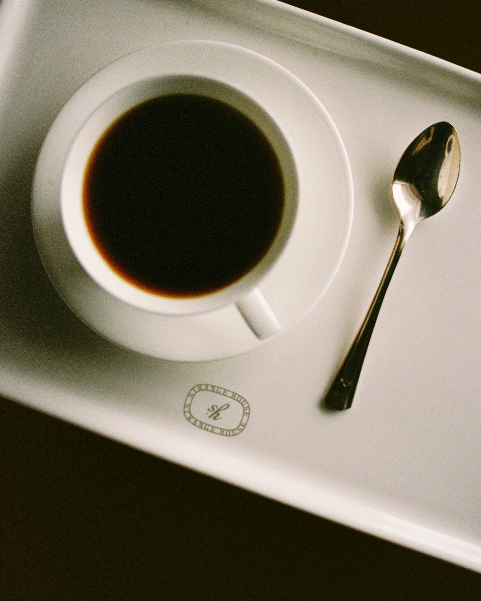 Overhead shot of a white coffee cup filled with dark coffee on a saucer next to a silver spoon on a white tray