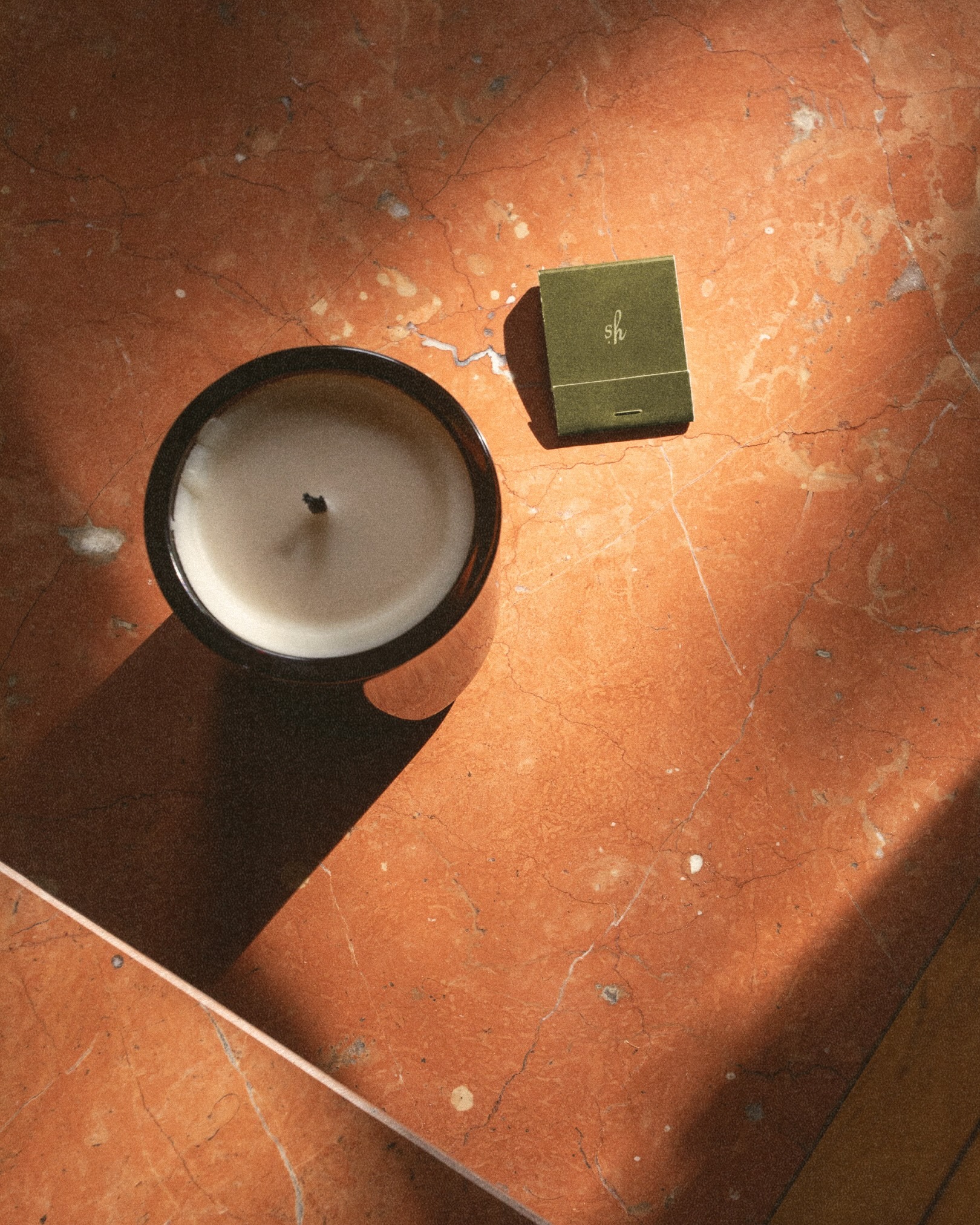 Overhead view of a black scented candle in a jar and a green matchbox resting on a warm orange marble surface with harsh sunlight