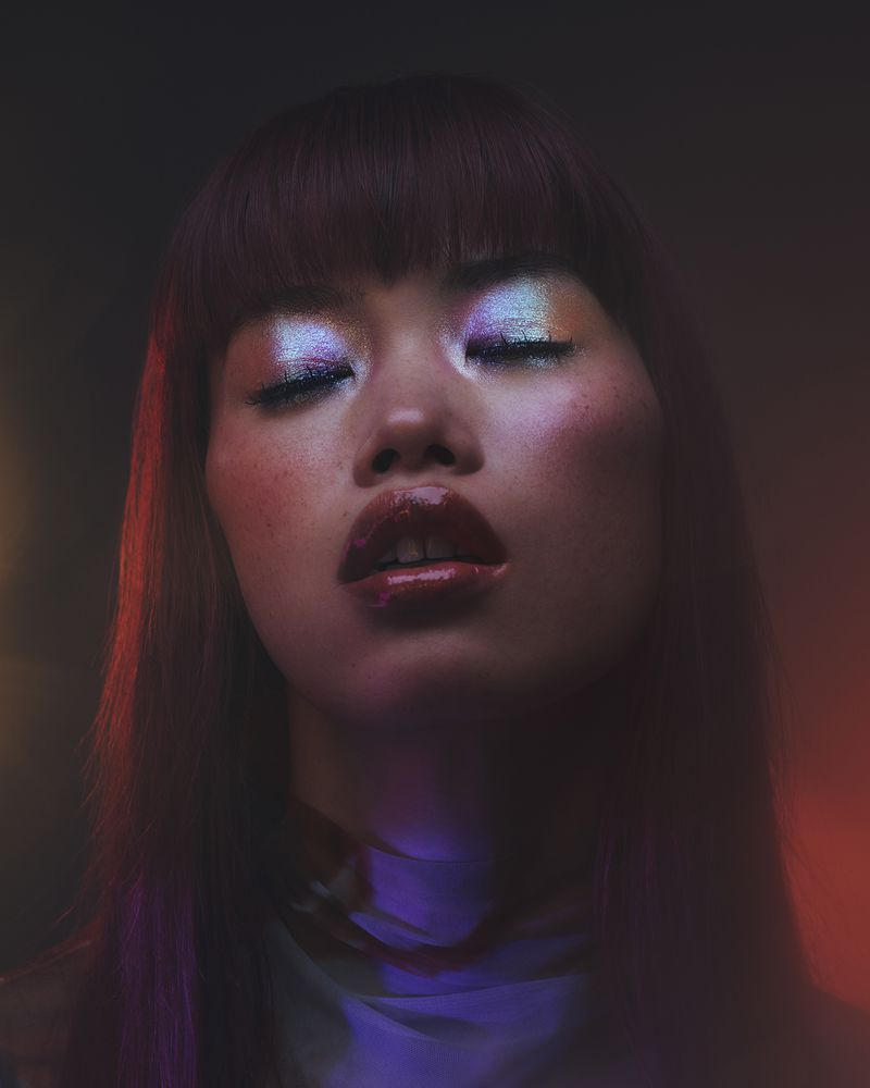 Close-up portrait of a model with iridescent metallic eyeshadow, glossy red lips, bangs, and colorful studio lighting.