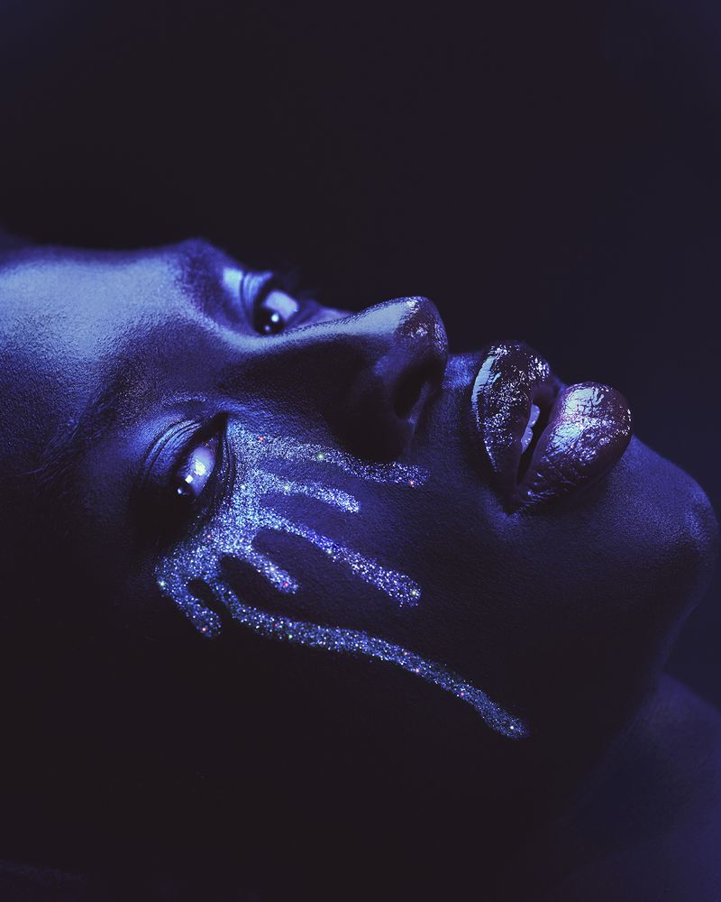 Close-up profile of a person with blue-toned lighting and sparkling glitter makeup dripping down the cheek from the eye.
