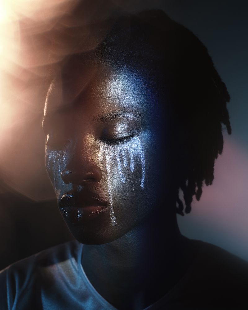 Close-up portrait of a Black woman with shimmering silver glitter tears on her face and dramatic blue and warm lighting.