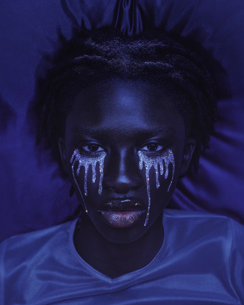 Close-up portrait of a Black woman with silver glitter tears makeup under blue lighting