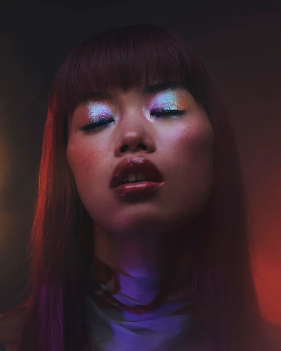 Close-up portrait of a model with iridescent metallic eyeshadow, glossy red lips, bangs, and colorful studio lighting.