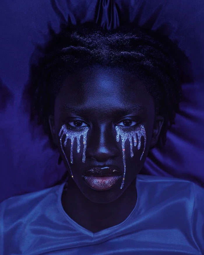 Close-up portrait of a Black woman with silver glitter tears makeup under blue lighting