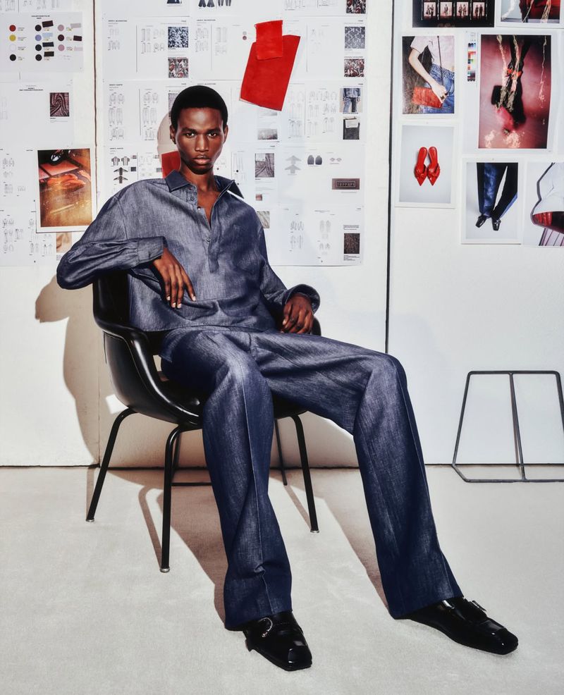 Adult male model posing in denim matching set in a photo studio environment with papers and images on the wall behind.