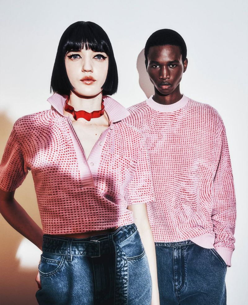 Two fashion models in a studio setting wearing matching pink tops and denim pants, posing in front of a neutral background with soft lighting.