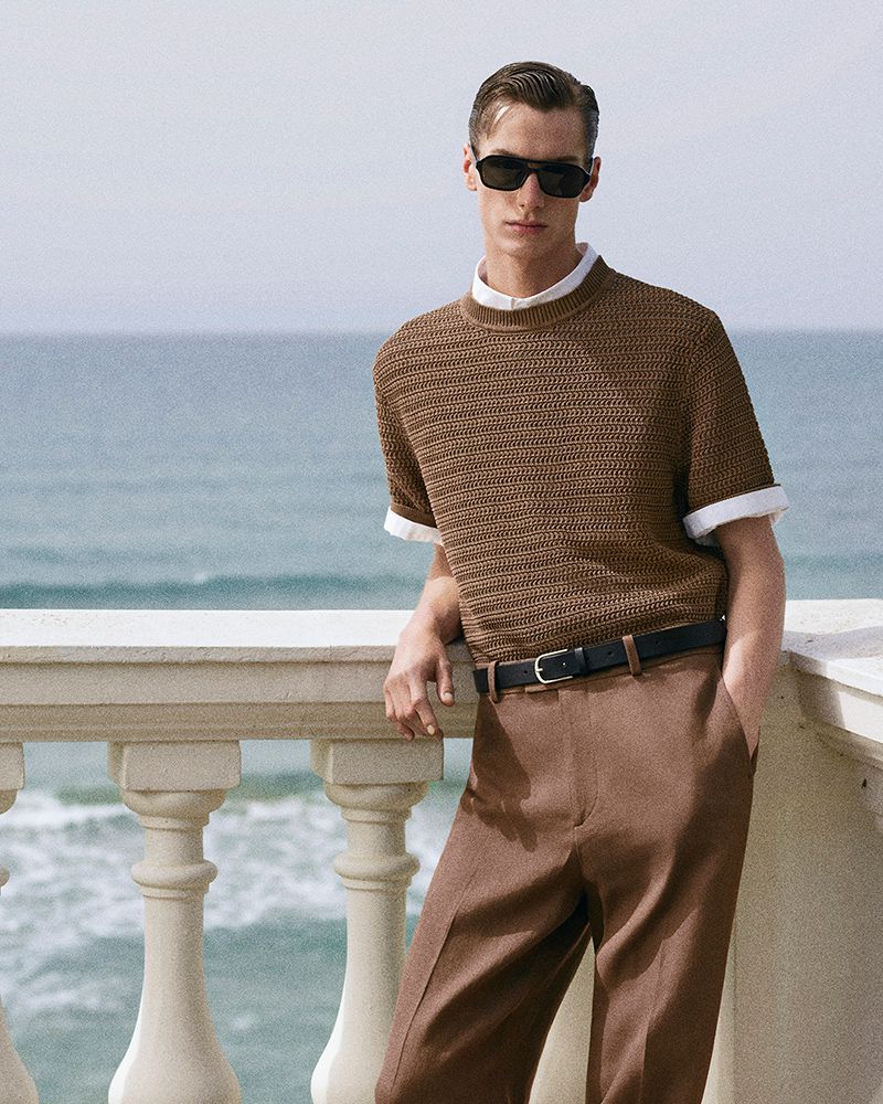 Model Leonard Kipker wearing a brown knit sweater and sunglasses leaning on a stone balustrade overlooking the ocean