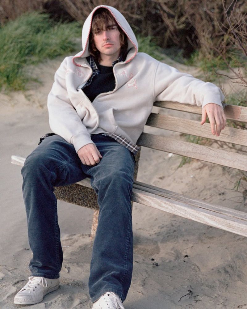 Gene Gallagher wearing a white hoodie and dark jeans sitting on a wooden bench outdoors in a sandy area for Madhappy.