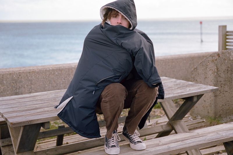 Person wrapped in a hooded changing robe sitting on a picnic table near the beach with the ocean and horizon in the background.