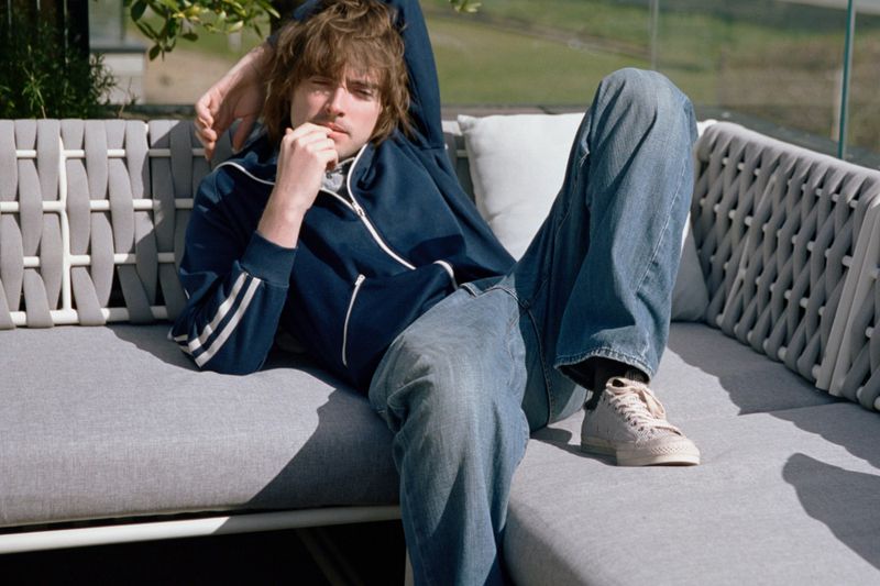 A young man reclining on a modern grey outdoor sofa, wearing a blue zip-up track jacket, blue jeans, and canvas sneakers.