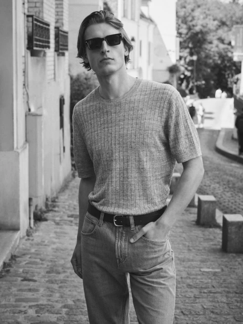 Portrait of a young adult man wearing sunglasses, a short-sleeve knit shirt, and jeans, posing on a cobblestone street with buildings.