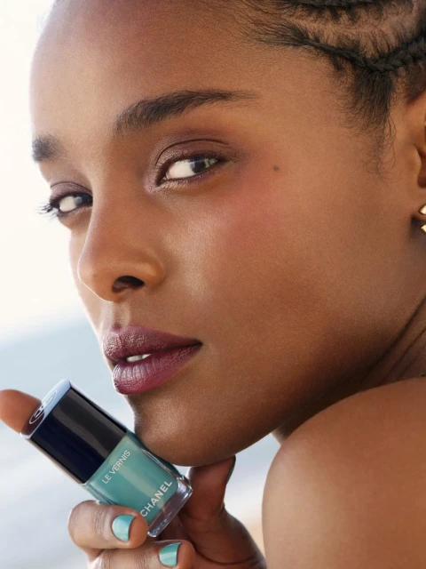 A close up of a Black female model holding a bottle of Chanel Le Vernis nail polish with turquoise manicure matching the bottle's hue.