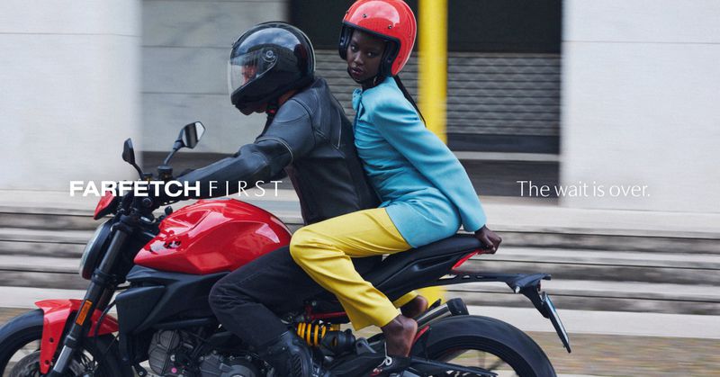 Two models riding a red motorcycle outdoors, wearing helmets, a black leather jacket, blue jacket, and yellow trousers.