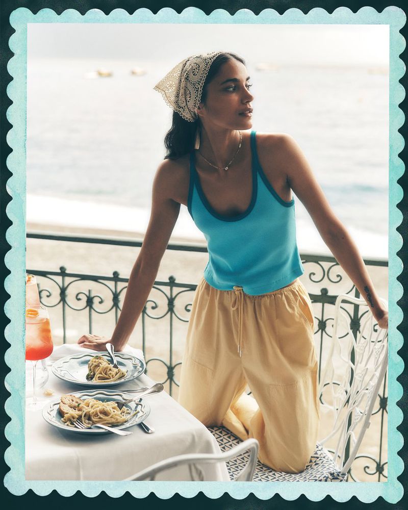 Model in blue tank top and yellow trousers kneeling by a dining table on a balcony overlooking the sea