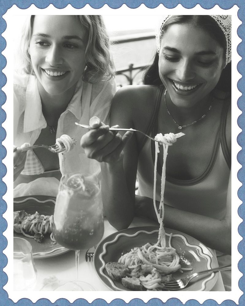 Two smiling models dining together, lifting pasta on forks with plates of food and a drink in the foreground