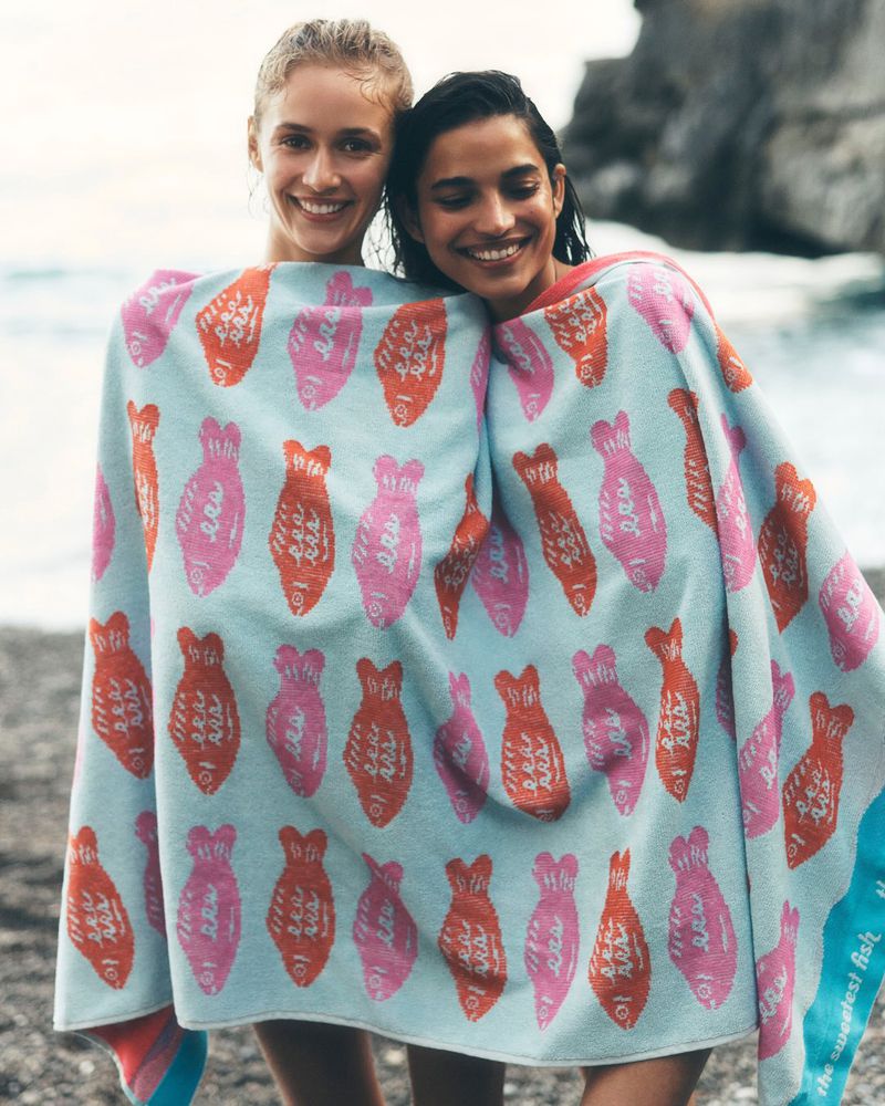 Two female models wrapped in a light blue patterned beach towel smiling together outdoors