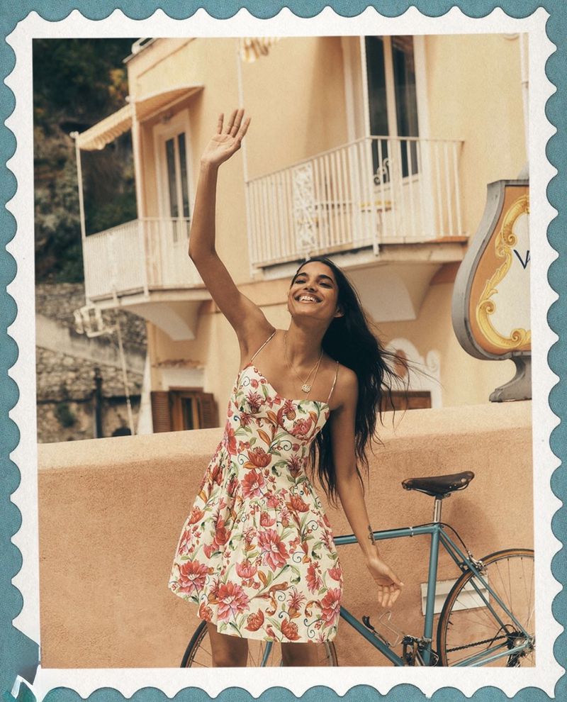 A cheerful young woman in a floral summer dress waves, standing next to a blue bicycle, with sunlit architecture in the background.