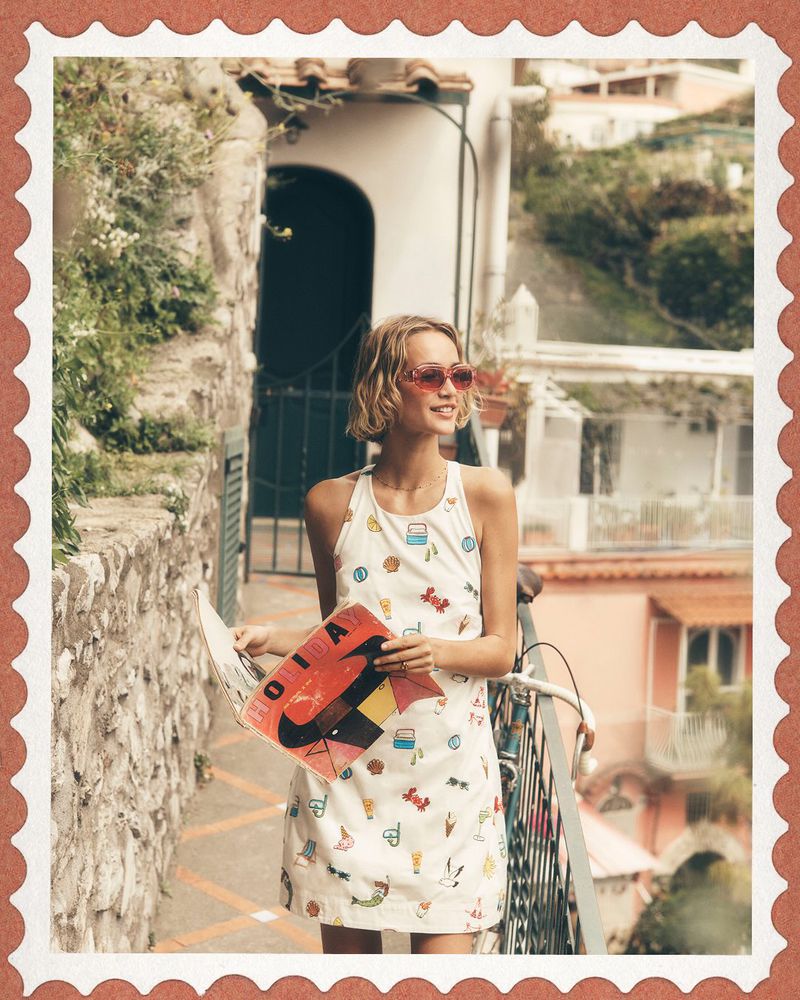 Model wearing patterned dress and pink sunglasses holding a magazine outdoors near a Mediterranean villa