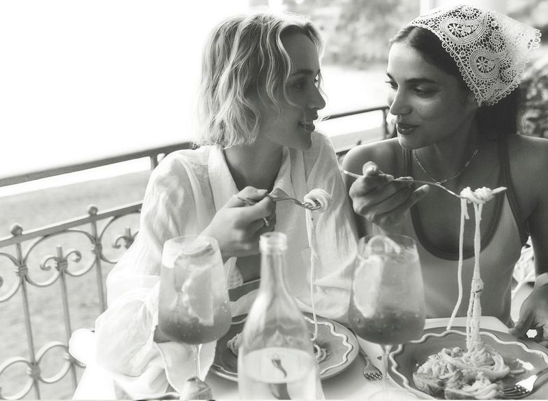 Two women dining outdoors at a balcony table eating pasta and drinks for Anthropologie Summer Said So campaign.