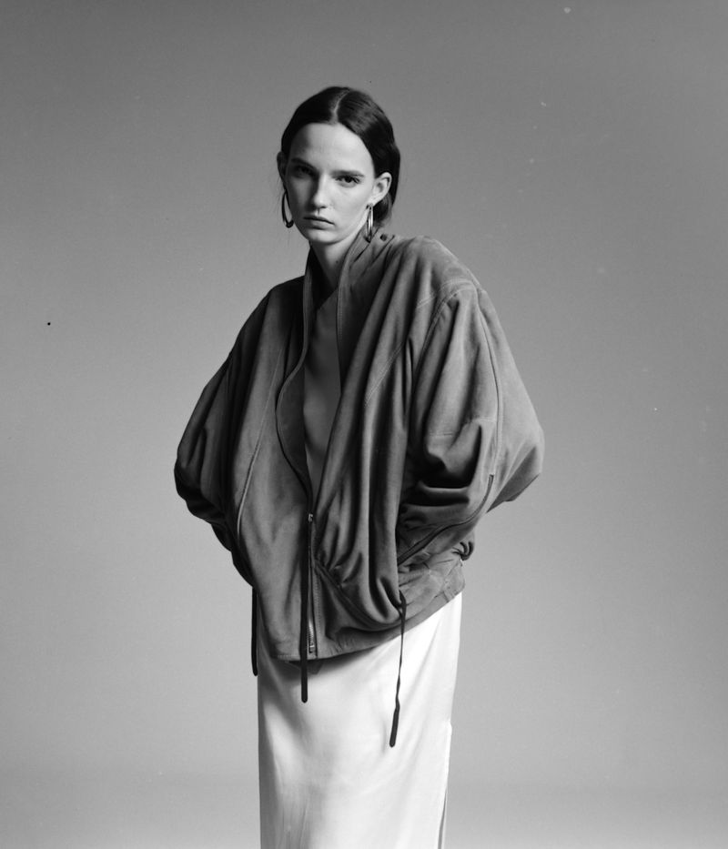 Monochrome portrait of a woman in a bomber jacket and skirt against a light gray backdrop, showcasing fashion and editorial style.