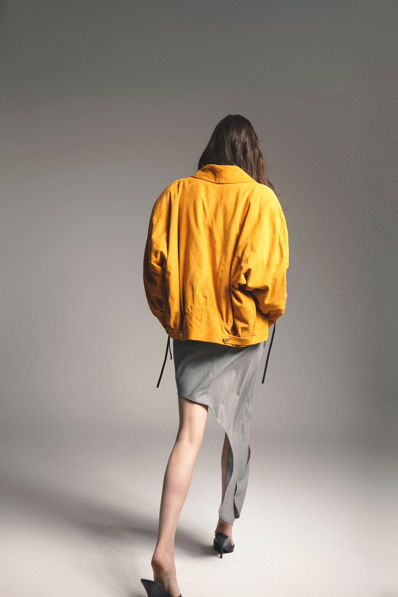 Model photographed from behind wearing a mustard yellow jacket and an asymmetrical grey skirt on a grey seamless background